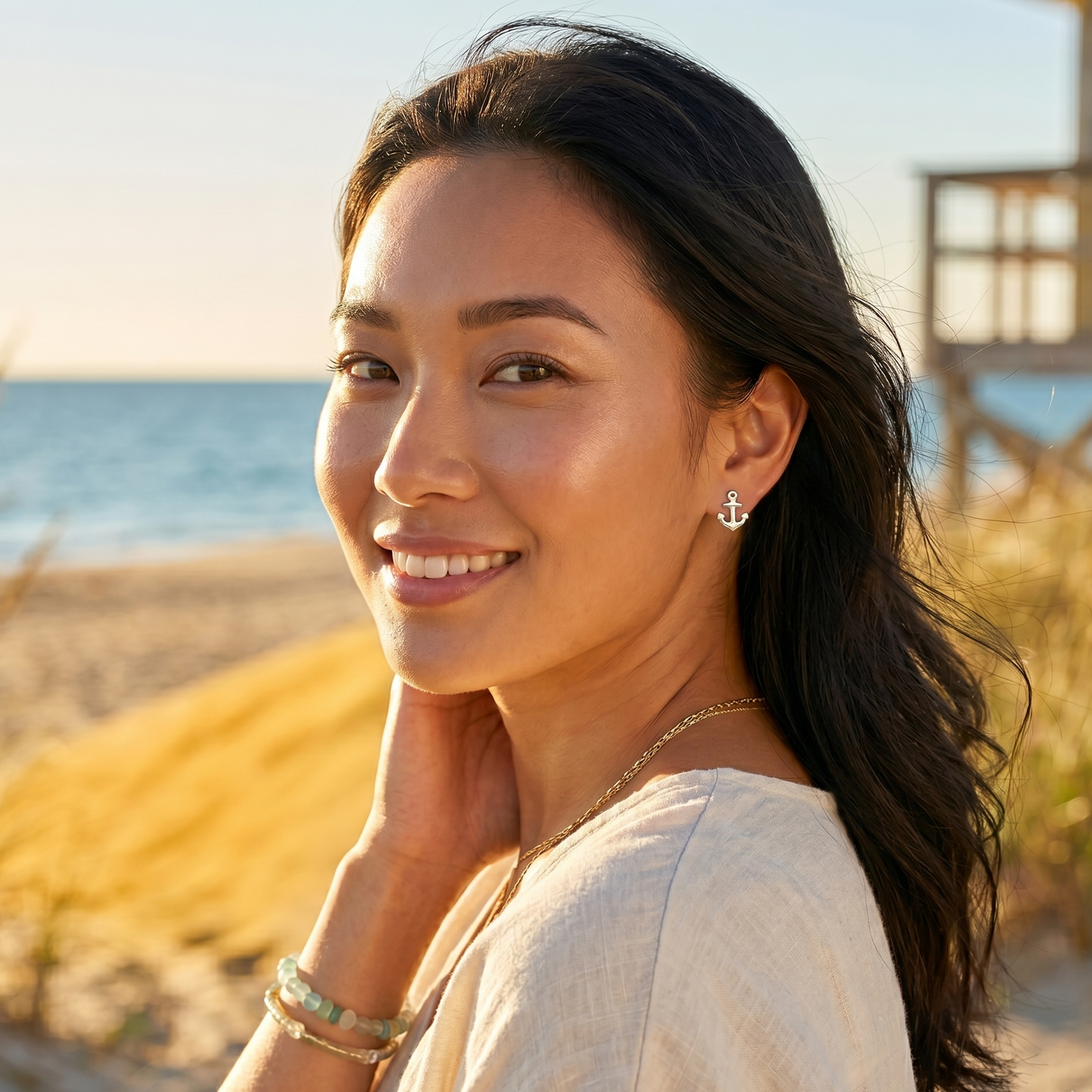 Woman standing on a beach with ocean view, wearing a light-colored top and jewelry.