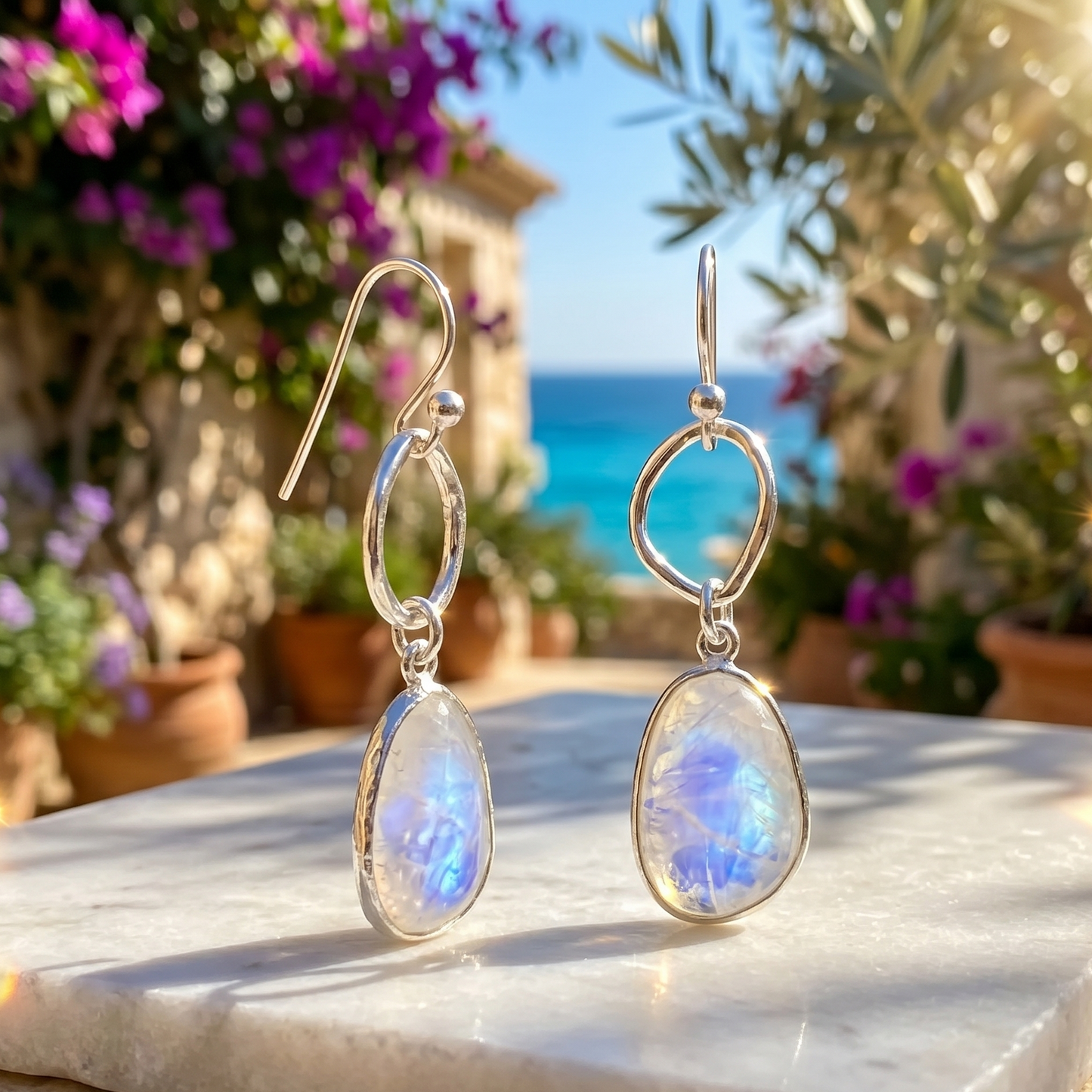 Pair of silver earrings with rainbow moonstone drops on a marble surface with a scenic background.