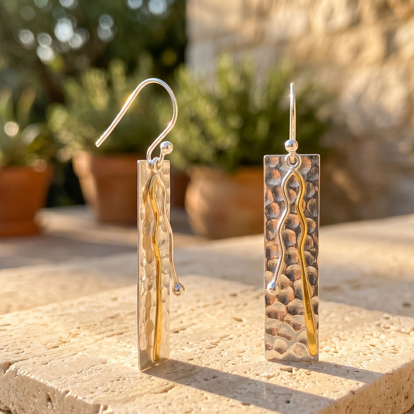 Pair of gold and silver earrings on a stone surface with plants in the background