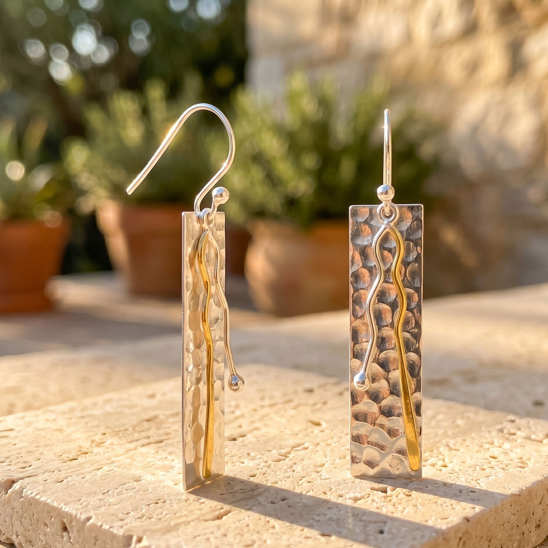 Pair of gold and silver earrings on a stone surface with plants in the background