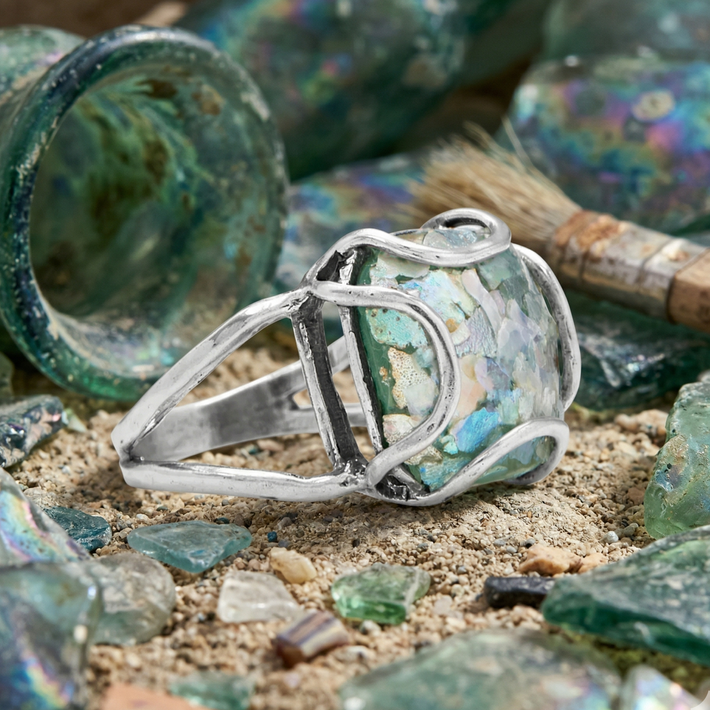 Silver ring with embedded glass碎片 on a sandy surface with broken glass pieces.